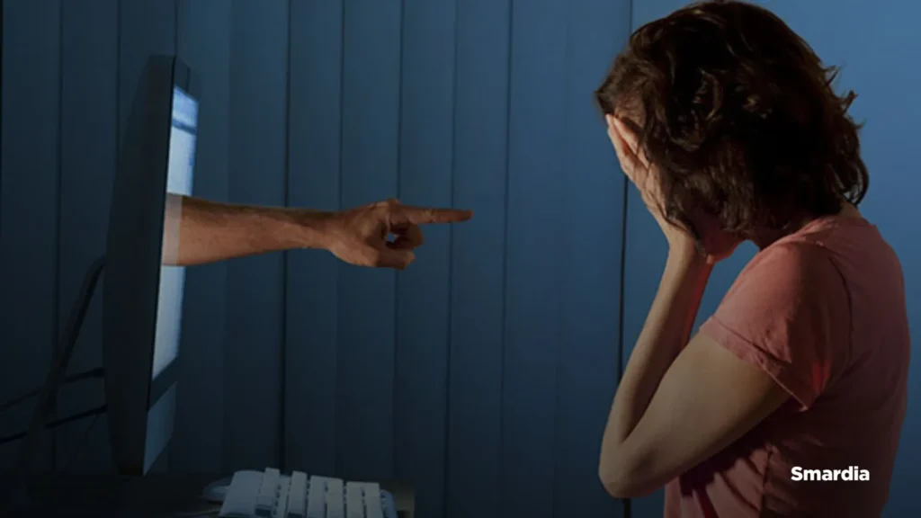 On voit un écran d’ordinateur qui pointe du doigt une jeune femme. Elle se cache le visage avec ses mains, car elle pleure.
